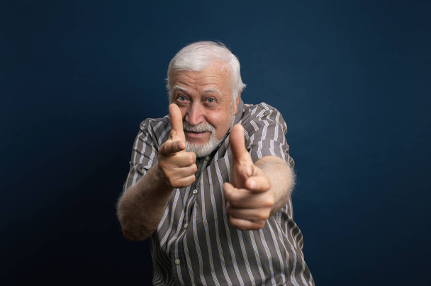 Guapo anciano enérgico, carismático hombre de pelo gris entrecerrando los ojos y mostrando los dedos índice delante de él, emociones, retrato de media longitud, fondo azul - Foto, imagen