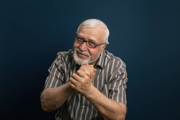 Guapo anciano enérgico, carismático hombre de pelo gris en gafas, dobló sus manos, emociones en el marco, retrato de media longitud, fondo azul - Foto, imagen