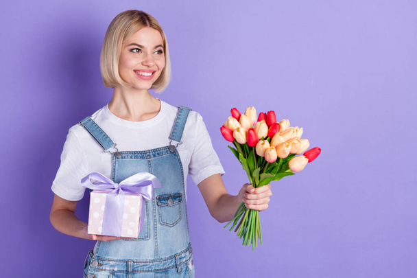 Photo of nice optimistic short hairdo blond lady hold flowers present look empty space wear white t-shirt isolated on violet background - Fotografie, Obrázek