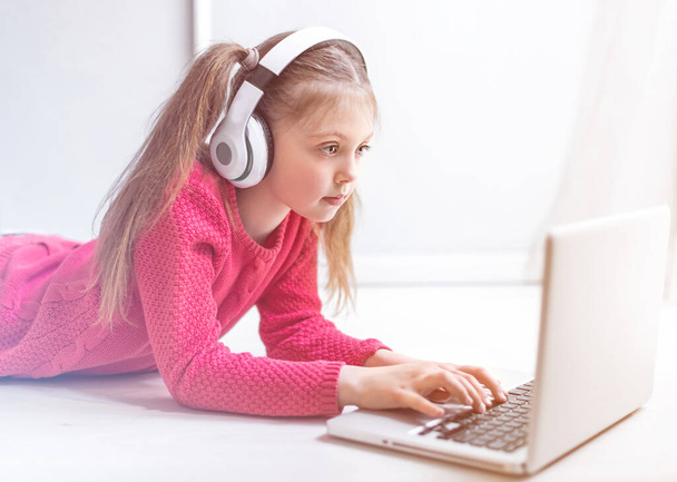 Cute little girl in headphones seriously using laptop at home for online education, home studying and online communication. Kids distance learning. Social distance. Stay at home. - Fotó, kép