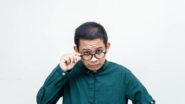 緑のシャツを着たアジア系マレー人の男に混乱した顔は、隔離された白い背景に眼鏡のペアを保持カメラを見て集中しています。彼の前で被験者を調べて. - 写真・画像
