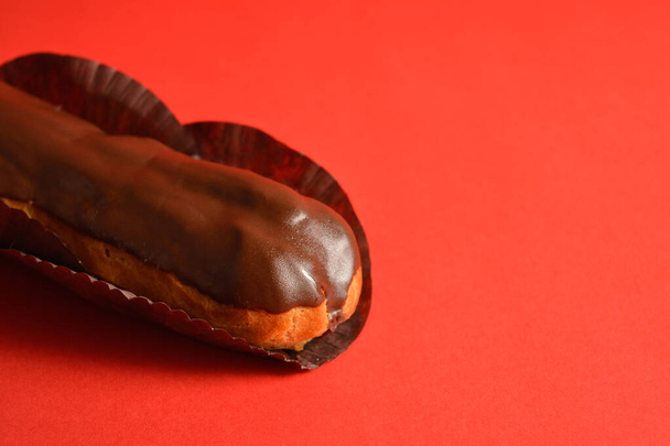 Close up to part of French dessert on red background. Eclair with custard filling and dark chocolate on top. Copy space image creative concept. Sweet menu object - Photo, Image