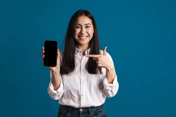 Счастливая красивая азиатская девушка показывая и указывая пальцем на смартфон изолированы на синем фоне - Фото, изображение