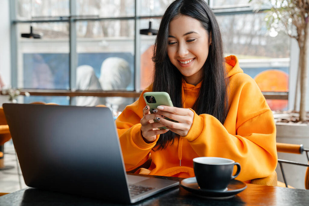 Smiling asian woman using mobile phone while working with laptop in cafe - Photo, Image