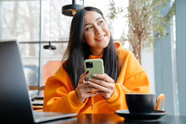 Smiling asian woman using mobile phone while working with laptop in cafe - Photo, Image