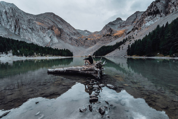 A vertical shot of a mountainous lake and a male sitting on wood in water - Foto, immagini
