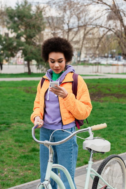 Mujer afroamericana joven usando teléfono celular mientras está parada cerca de la bicicleta al aire libre  - Foto, imagen