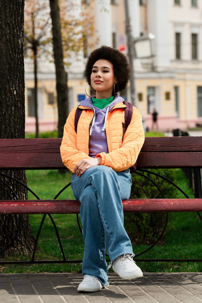 African american woman in jacket sitting on bench on urban street  - Photo, Image