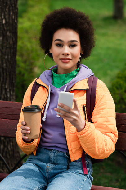Femme afro-américaine avec téléphone portable et tasse en papier regardant la caméra sur le banc dans le parc  - Photo, image