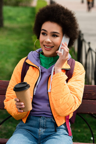 Pretty african american woman with takeaway beverage talking on mobile phone on bench outdoors  - Photo, Image