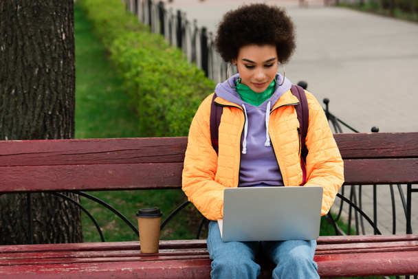 Femme afro-américaine avec sac à dos en utilisant un ordinateur portable près du café pour aller sur le banc  - Photo, image