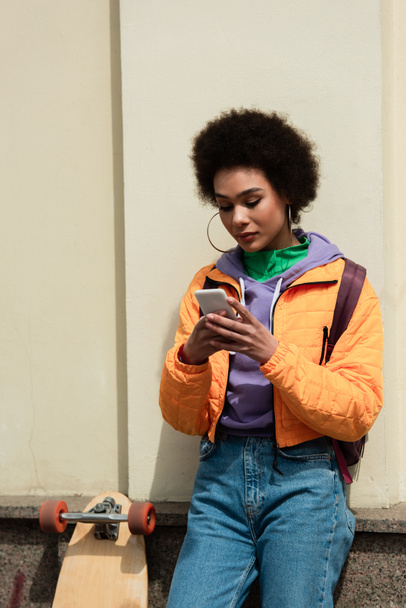 African american woman using smartphone near building and longboard outdoors  - 写真・画像