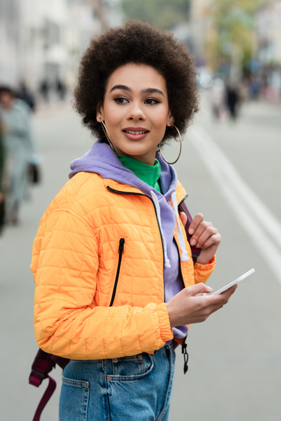 笑顔アフリカ系アメリカ人女性とともに携帯電話やバックパックを離れて屋外で見る  - 写真・画像