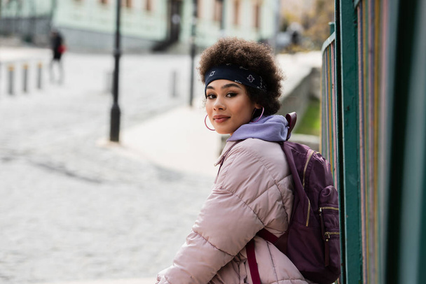 Młoda Afroamerykanka z plecakiem patrząca na kamerę w pobliżu ogrodzenia na ulicy miejskiej  - Zdjęcie, obraz