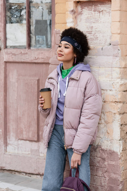 Pretty african american woman with backpack holding coffee to go near old building  - Foto, Imagen