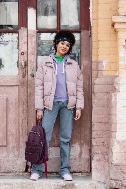 African american woman with backpack standing near old building with wooden door  - Фото, изображение