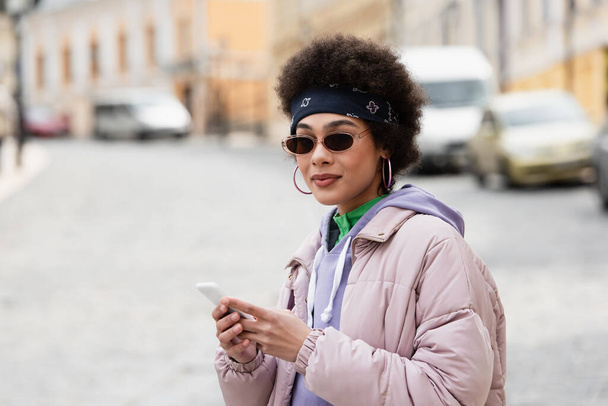 Afro-Amerikaanse vrouw in zonnebril en bandana houden mobiele telefoon op stedelijke straat  - Foto, afbeelding