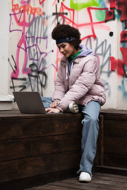 Afro-Amerikaanse telewerker met behulp van laptop op bank in de buurt van graffiti buiten  - Foto, afbeelding