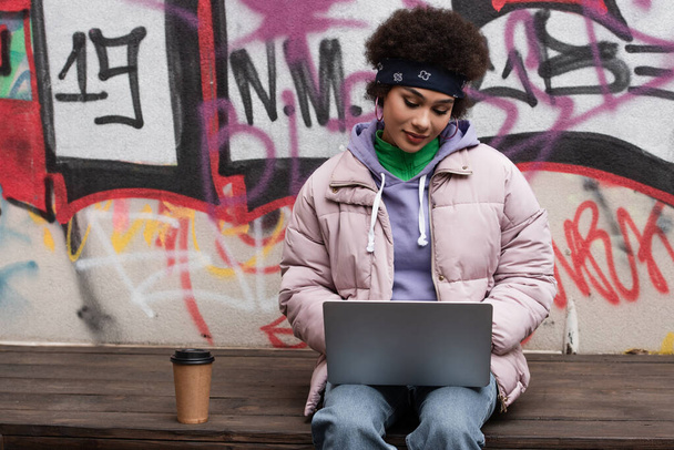 Afrykański freelancer w kurtce przy laptopie przy drinku na wynos na ławce i graffiti na ścianie  - Zdjęcie, obraz