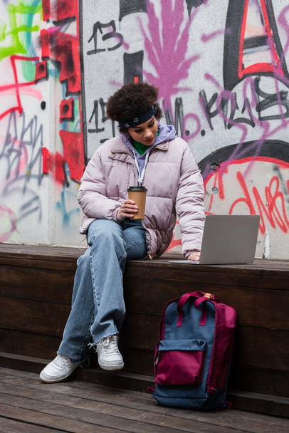 Africano americano mulher com café para ir usando laptop no banco perto de mochila e graffiti  - Foto, Imagem