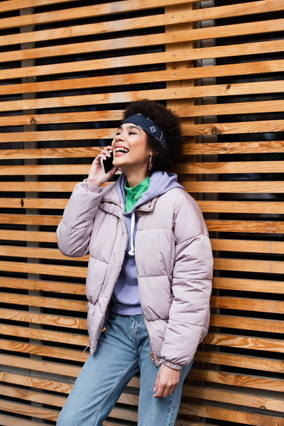 バンダナとジャケットの幸せなアフリカ系アメリカ人女性は木製フェンスの近くのスマートフォンで話しています  - 写真・画像