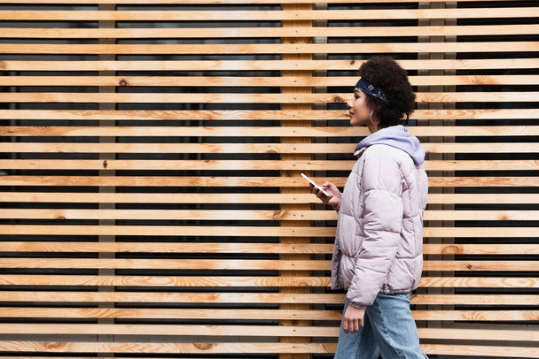 Seitenansicht einer afrikanisch-amerikanischen Frau, die mit ihrem Smartphone in der Nähe eines Holzzaunes läuft  - Foto, Bild
