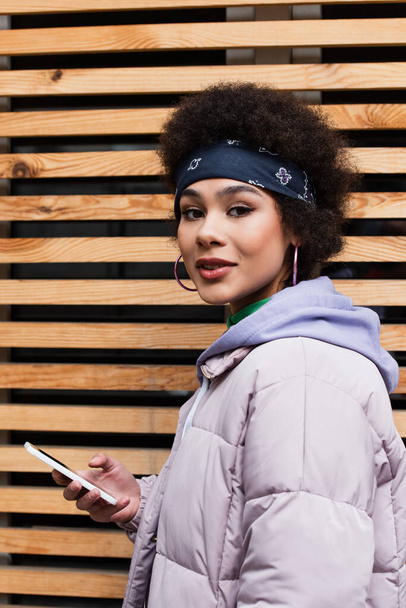 African american woman in jacket holding mobile phone and looking at camera outdoors  - Valokuva, kuva