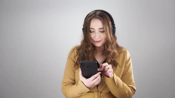 Atraktivní žena ve sluchátkách vybrat hudbu v telefonu a tance stojící na bílém pozadí - Záběry, video
