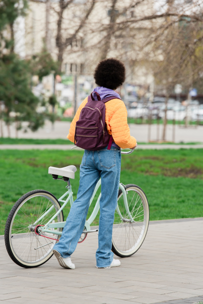 Back view of african american woman with backpack holding bicycle and walking outdoors   - Valokuva, kuva