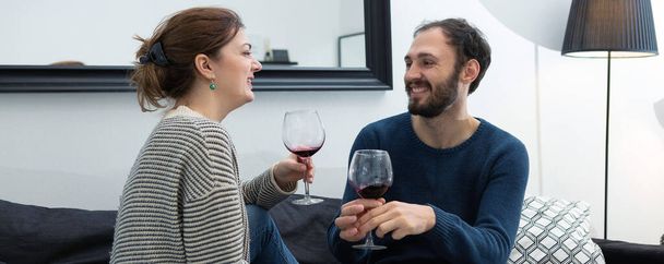 Νεαρό και ευτυχισμένο ζευγάρι πίνοντας κρασί και χαλαρώνοντας στο σπίτι - Φωτογραφία, εικόνα
