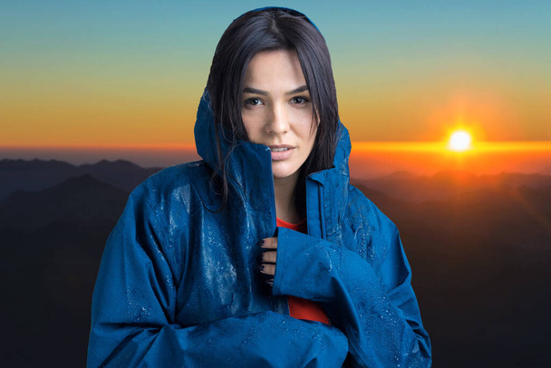 Retrato de una niña sonriente vestida con impermeable azul en gotas posando con capucha sobre el fondo de la puesta de sol en las montañas. - Foto, imagen