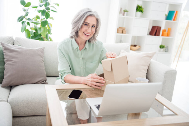 Photo of pretty cheerful person sit on couch hands open paper package beaming smile look camera indoors - Photo, image