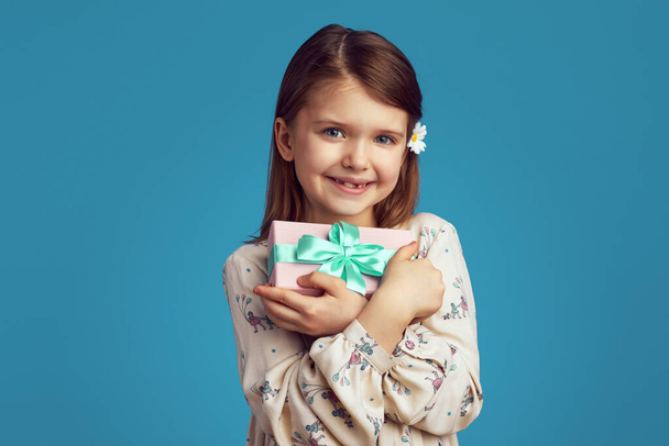 Genç kız gülümsüyor ve mavi arka plana sarılmış hediye kutusunu kucaklıyor. - Fotoğraf, Görsel