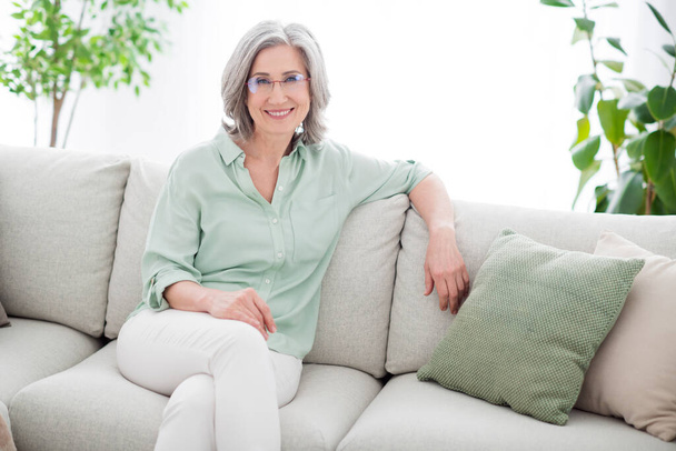 Photo de joyeuse vieille grand-mère retraitée assis canapé porter des lunettes sourire à l'intérieur de la maison maison - Photo, image