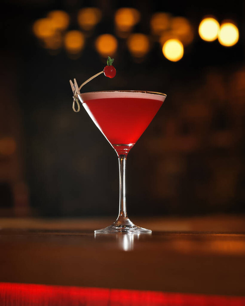Rode alcoholische cocktail versierd met bessen aan houten tafel. Transparant glas van alcoholische drank geïsoleerd op zwarte achtergrond met bokeh achtergrondverlichting - Foto, afbeelding