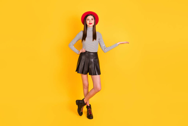 Φωτογραφία από αξιολάτρευτο λαμπερό νεαρή γυναίκα φορούν ριγέ πουκάμισο καπέλο εκμετάλλευση βραχίονα κενό χώρο απομονωμένο κίτρινο χρώμα φόντο - Φωτογραφία, εικόνα