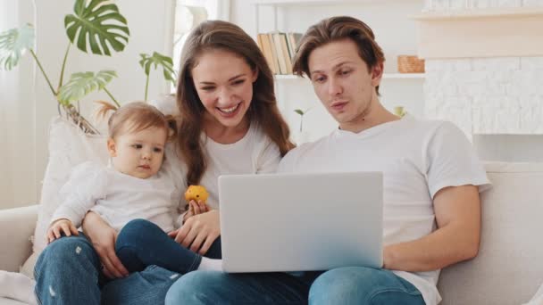 Kaukaska rodzina młodzi rodzice i mała córeczka dziewczynka siedzi na kanapie w domu kupując towary online i w sklepie internetowym za pomocą laptopa przeglądarki sieci wybrać opiekunka na stronie - Materiał filmowy, wideo