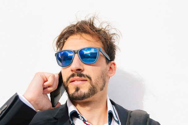 Portrait of an adult businessman with blue sunglasses, using his mobile phone, with white outdoor background and copy space. - Foto, imagen