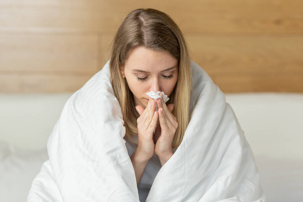 young sick woman sitting on the bed in the bedroom wrapped in a blanket with a runny blows his nose into a napkin. Cold female or girl or flu at home. Close-up portrait Attractive young blonde - Fotoğraf, Görsel