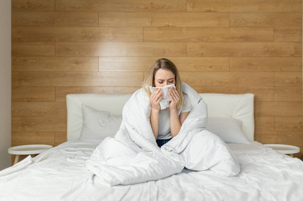 young sick woman sitting on the bed in the bedroom wrapped in a blanket with a runny blows his nose into a napkin. Cold female or girl or flu at home. Close-up portrait Attractive young blonde - Foto, Imagem