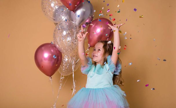 fun little girl stands with balloons and plays with colorful confetti on a beige background with space for text - Photo, Image