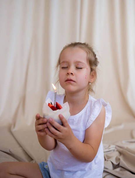 μικρό κορίτσι γενεθλίων κρατά ένα cupcake με ένα κερί και κάνει μια ευχή σε ένα μπεζ φόντο με μια θέση για κείμενο - Φωτογραφία, εικόνα