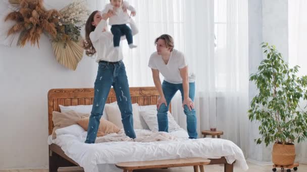 Kavkazský mladý rodina rodiče baví doma v ložnici stojící na posteli, silný dospělý matka maminka zvedání dítě dcera holčička do vzduchu házení vysoké dítě létání otec aplauds podporuje - Záběry, video