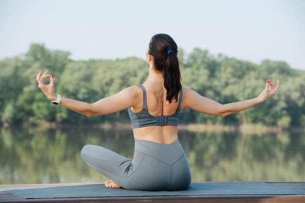 Sportive woman doing yoga outdoors in a beautiful spot on a side of the river. Trees reflecting in the water. She's meditating with hands outstretched to sides and fingers joined. - Foto, Imagen