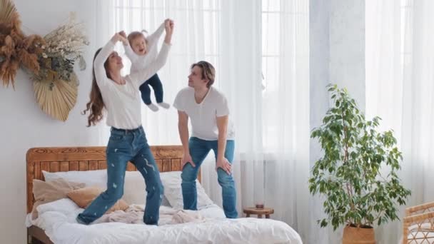 Young parents caucasian couple playing with little daughter child girl mother strong woman lifts baby in air holding hands doing acrobatic position flight caring man support, family weekend in bedroom - Footage, Video