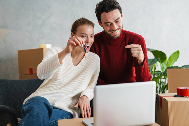 Smiling couple with laptop sitting on floor in new house, showing keys - 写真・画像