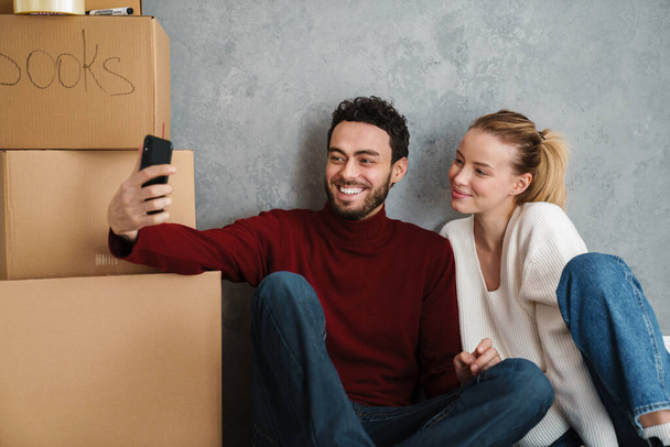 Bonito casal feliz tirando uma selfie em sua nova casa cercada de caixas - Foto, Imagem