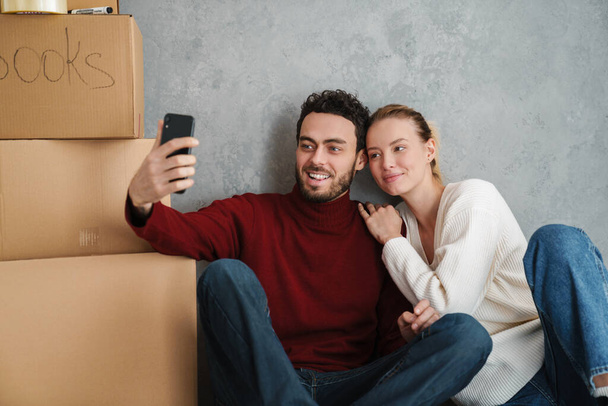 Cute happy couple taking a selfie in their new home surrounded with boxes - Zdjęcie, obraz