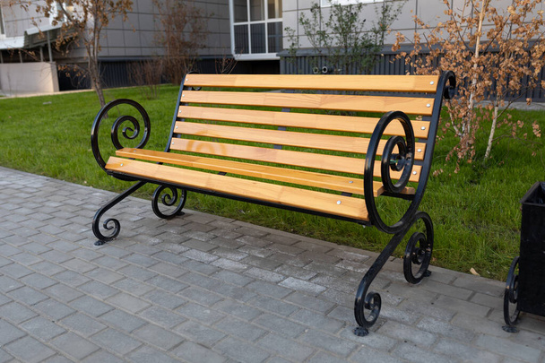 Classic wooden bench with metal armrests near an apartment building. Paving slabs under the bench. Improvement of public space in the urban environment. - Photo, Image