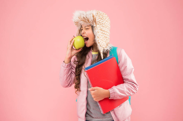 school break. small happy girl earflap hat eat apple. winter holiday and vacation. teen kid pink background. pupil daily life. Modern education. schoolgirl with books after lesson - Фото, зображення